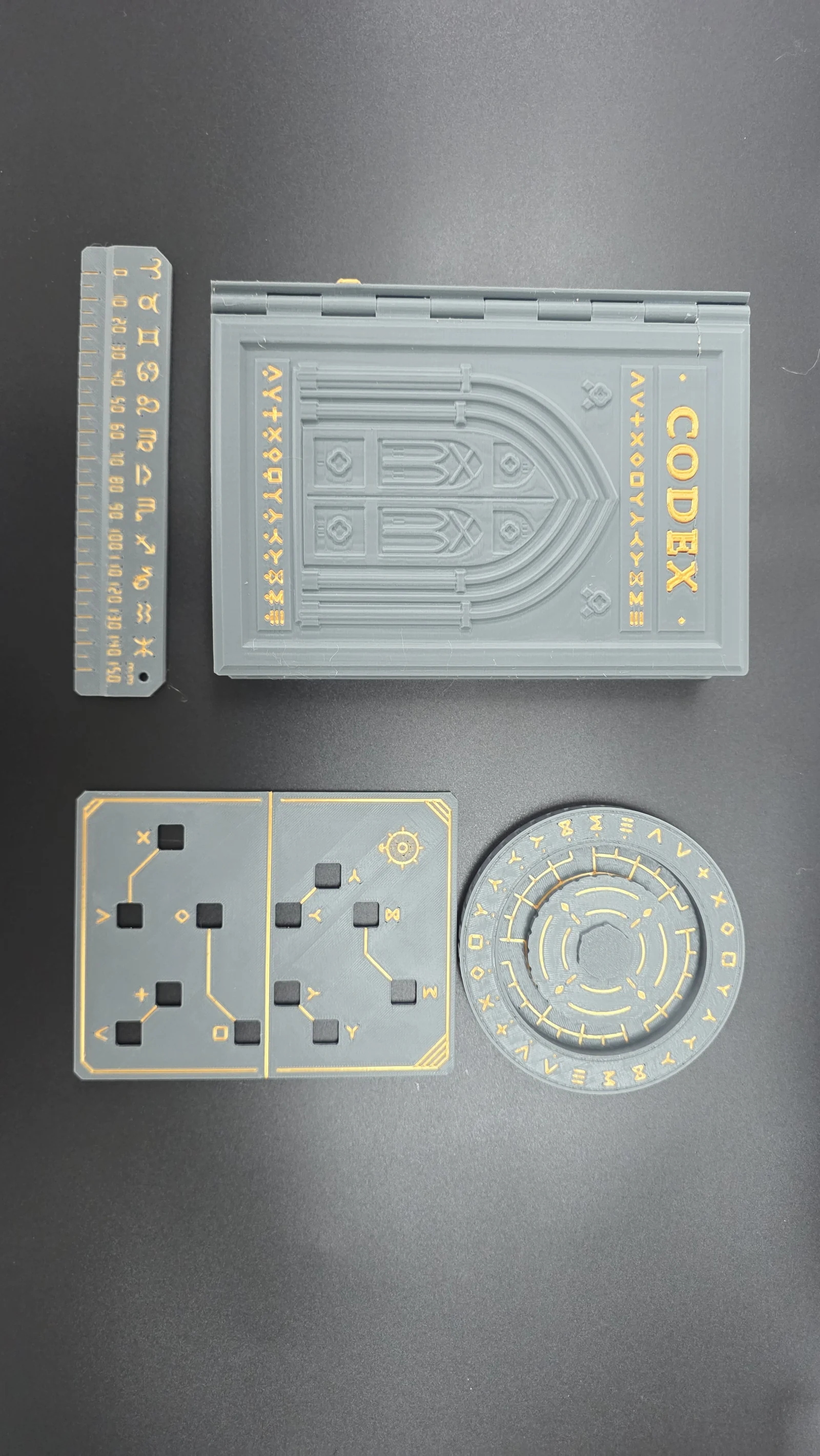 A grey and gold 3D-printed Codex, ruler, grille and wheel arranged on a dark tabletop.